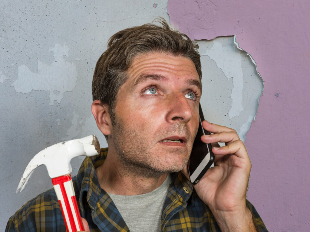 funny portrait of messy and frustrated man calling domestic insurance on mobile phone for repair mess he did with a hammer cracking the wall needing professional help of repairman